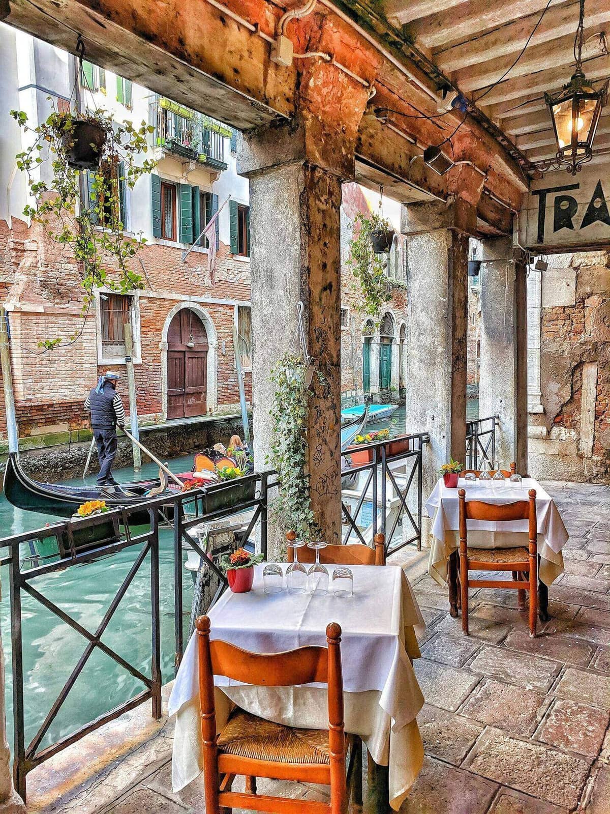 A working boat moves along a canal in central Venice next to a restaurant terrace, daily life.